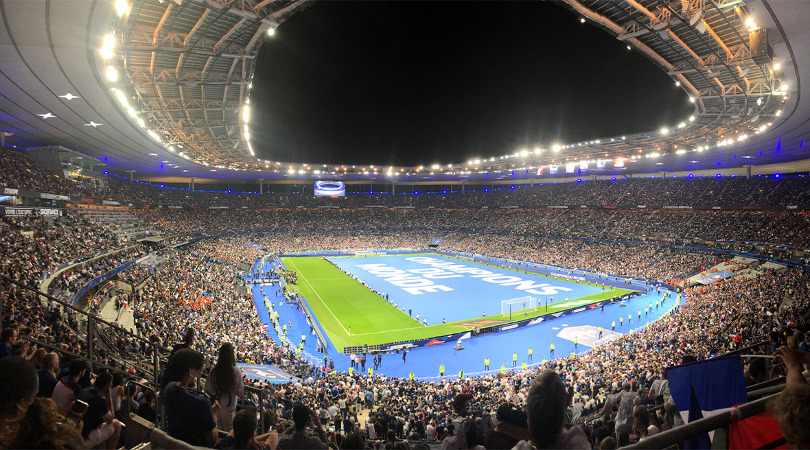 A large three tier stadium at night, with a capacity crowd, before a football match. The pitch is covered in a blue sheet with ‘Champions du Monde’ written on it